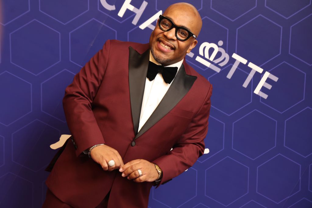 A smiling man wearing a red tuxedo jacket and bowtie stands in front of a purple backdrop with a "Charlotte" logo.