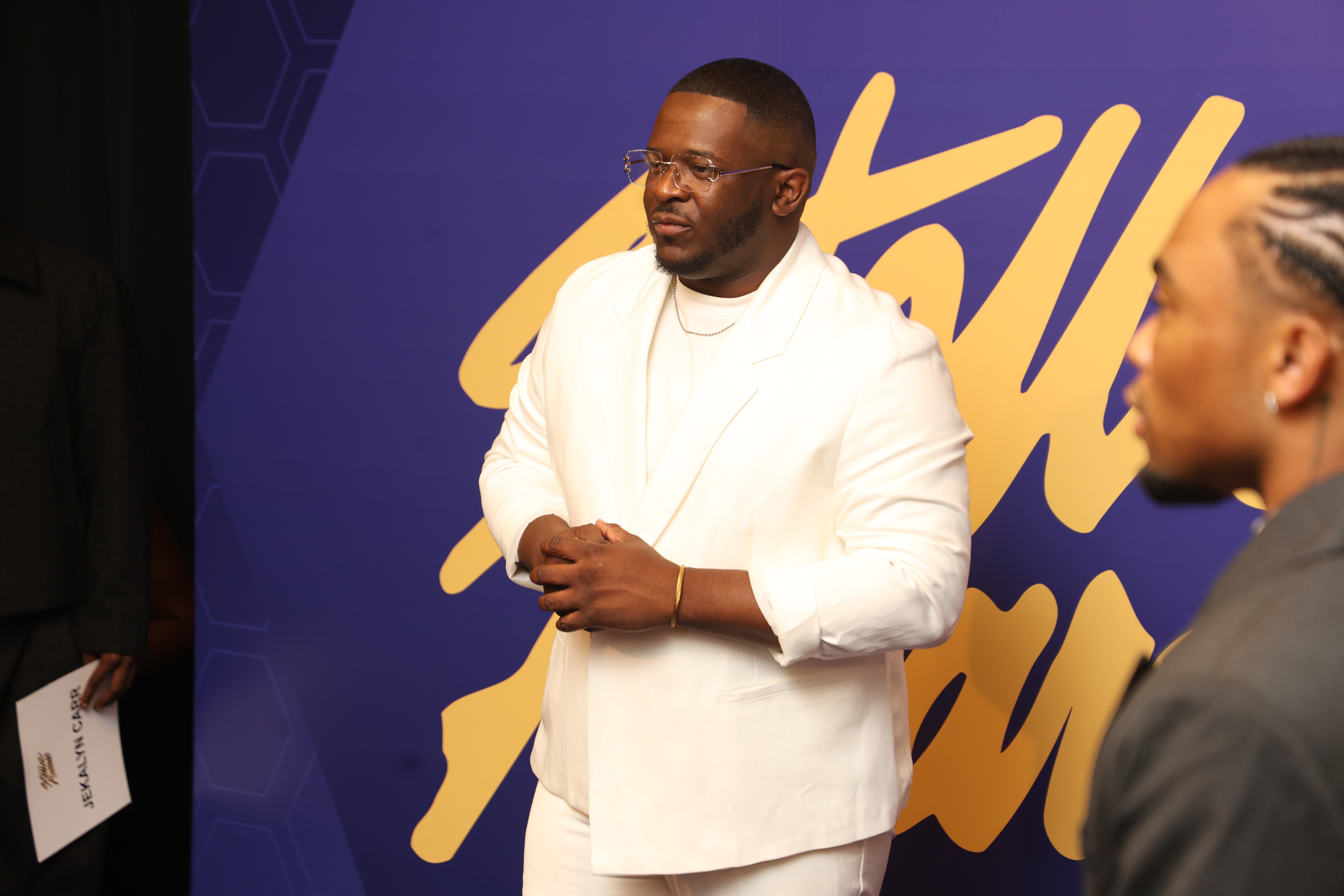 A man in a white suit standing on a stage with a purple and yellow background.