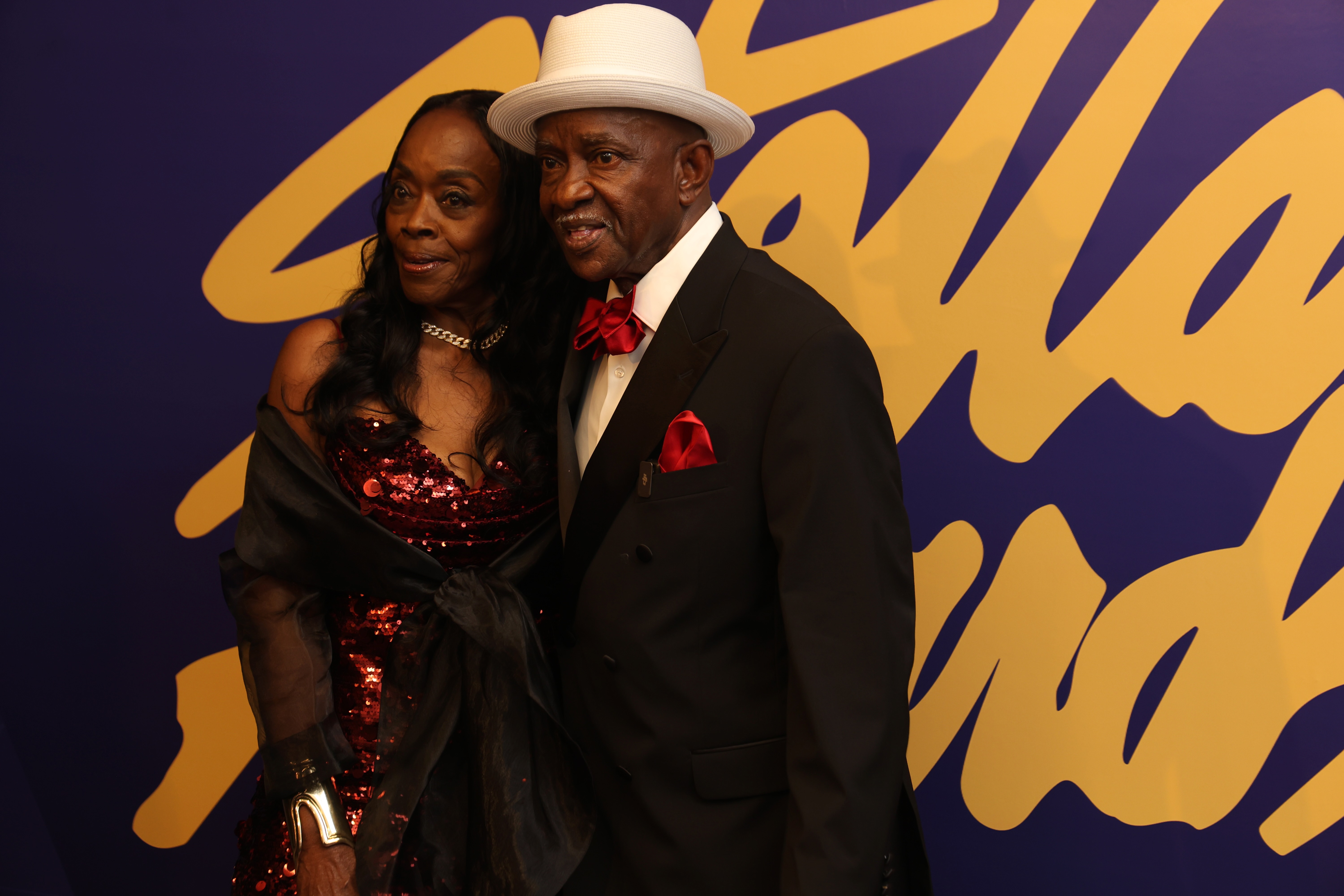 An older Black couple dressed formally in evening attire posing together in front of a vibrant abstract mural backdrop.