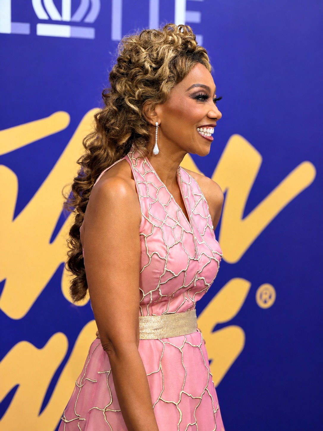 A smiling woman with curly brown hair wearing a pink and gold patterned dress posing in front of a blue and yellow background.