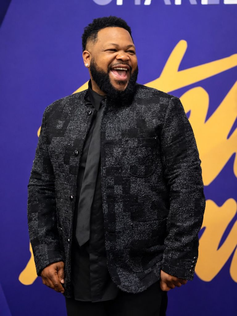 A smiling man with a beard wearing a black suit jacket stands in front of a purple and yellow background.